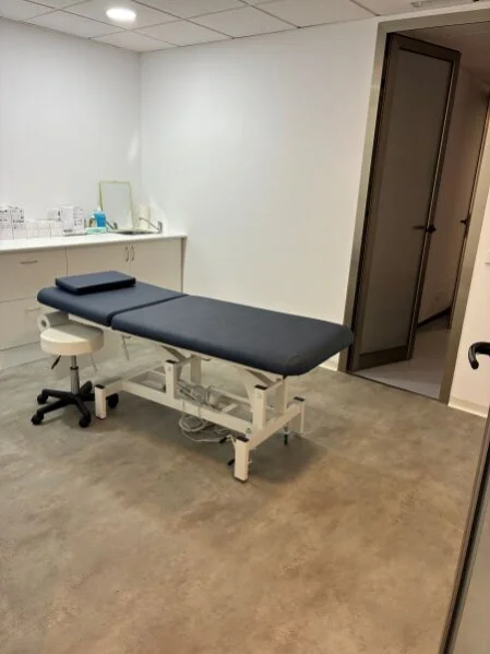 Eingriffsraum mit voll elektrischer, kompfortabel gepolsterter Behandlungsliege; Tür zum Umkleide- und Ruheraum mit Rettungsliege und Defibrillator.